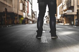 Person standing on an empty urban street, wearing jeans and black sneakers, with buildings in the background and a soft focus effect.