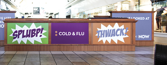 Promotional display at a retail location featuring vibrant comic-style graphics with expressions like "SPLUBB!" and "THWACK!" along with a central focus on "COLD & FLU" messaging, designed to attract attention to health products.