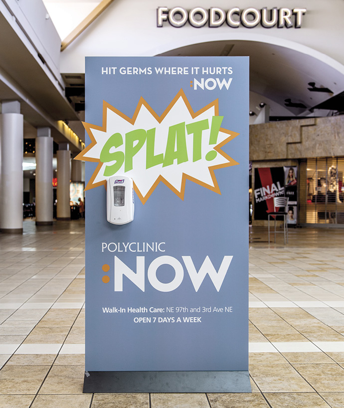 Signage promoting Polyclinic NOW's walk-in health care services, featuring a hand sanitizer dispenser and the slogan "Hit Germs Where It Hurts" with a vibrant "SPLAT!" graphic, located in a food court area. Open seven days a week at NE 97th and 3rd Ave NE.