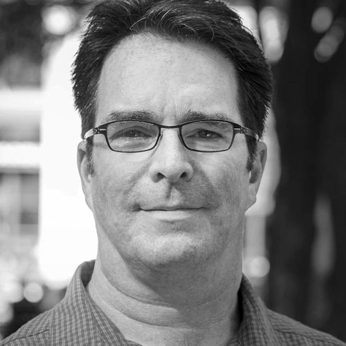 Middle-aged man with glasses, smiling confidently outdoors in a blurred urban setting.