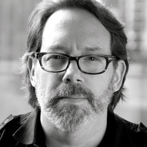 Black and white portrait of a middle-aged man with glasses and a beard, looking directly at the camera with a neutral expression. The background is softly blurred, emphasizing his face.