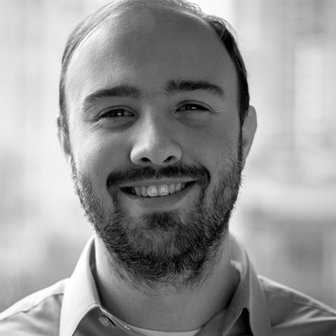 Smiling man with a beard wearing a light-colored shirt, captured in a black and white portrait against a blurred urban background.