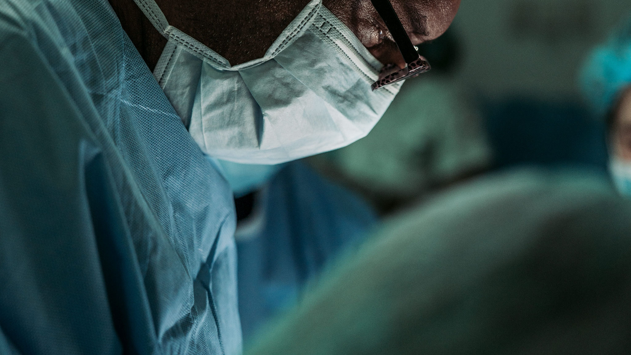Surgeon wearing a surgical mask and blue scrubs, focused on a medical procedure in a sterile environment.