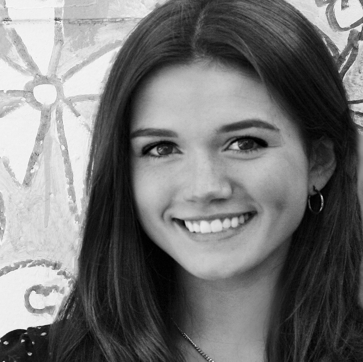 Smiling young woman with long brown hair and earrings, posed in front of a colorful painted backdrop, showcasing a joyful expression.