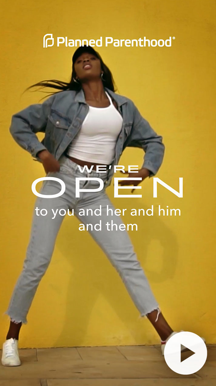 A confident model wearing a denim jacket and light jeans poses against a vibrant yellow background, with text stating "We're open to you and her and him and them," promoting inclusivity and diversity in fashion.