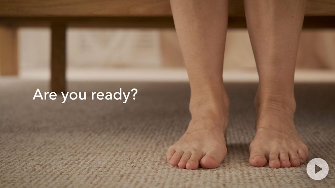Feet of a person standing on a carpet, with a wooden bed frame in the background, conveying a sense of readiness and anticipation.