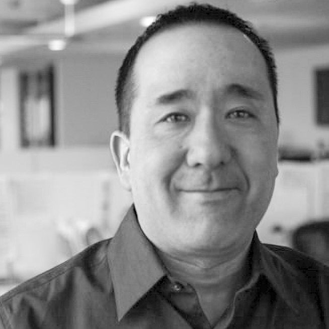Portrait of a smiling man in a black shirt, set in an office environment with blurred background elements, conveying a professional atmosphere.