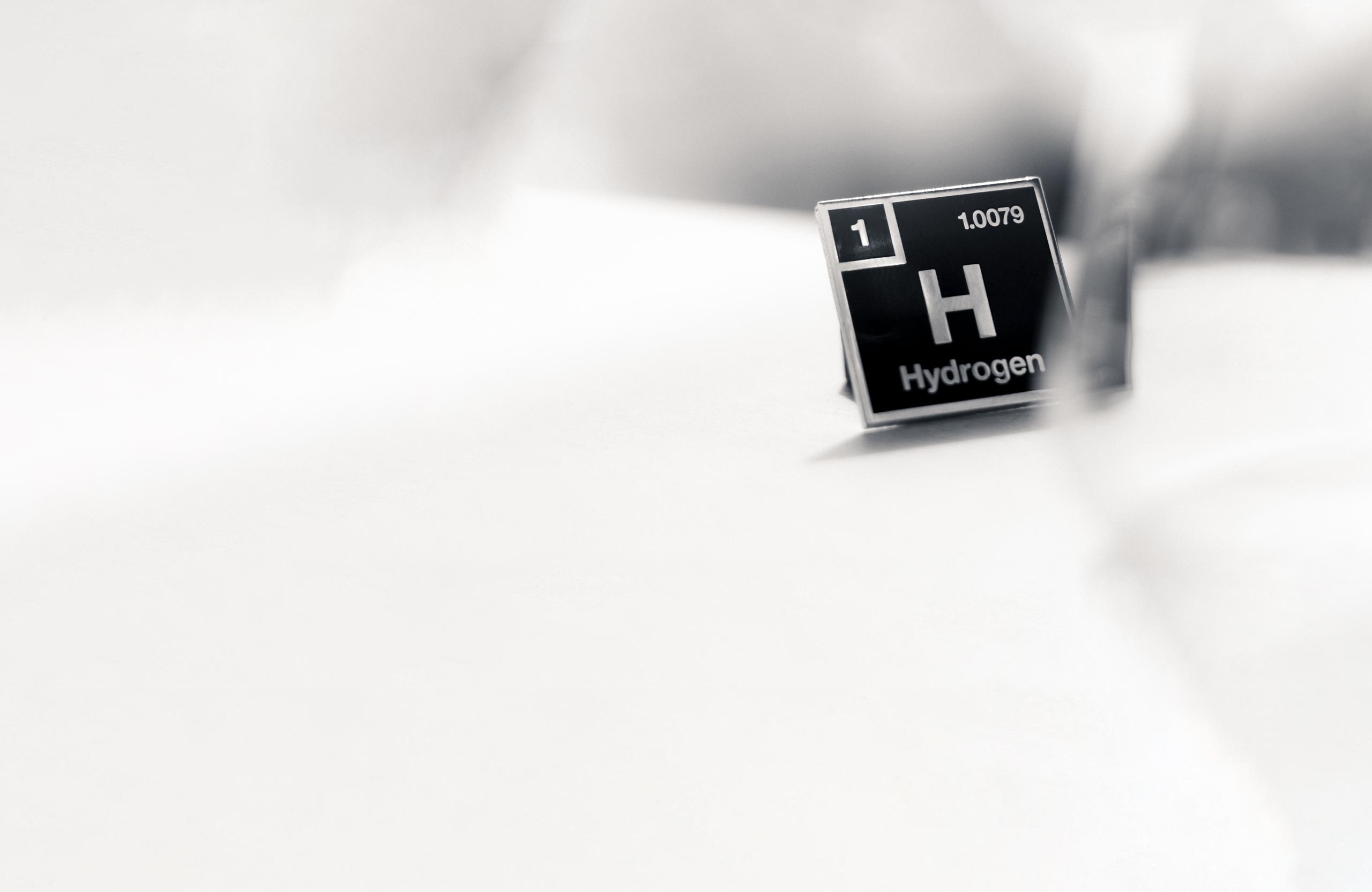 Close-up of a periodic table element card for hydrogen, showing the atomic number 1, symbol H, and the name "Hydrogen," with a blurred background.