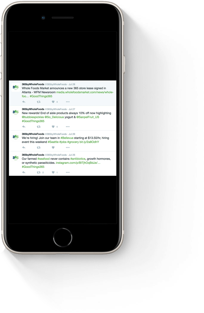 Twitter feed displayed on a smartphone screen featuring updates from 365 by Whole Foods, including announcements about a new store lease in Atlanta, product promotions, job openings in Bellevue, and information on their farmed seafood practices.