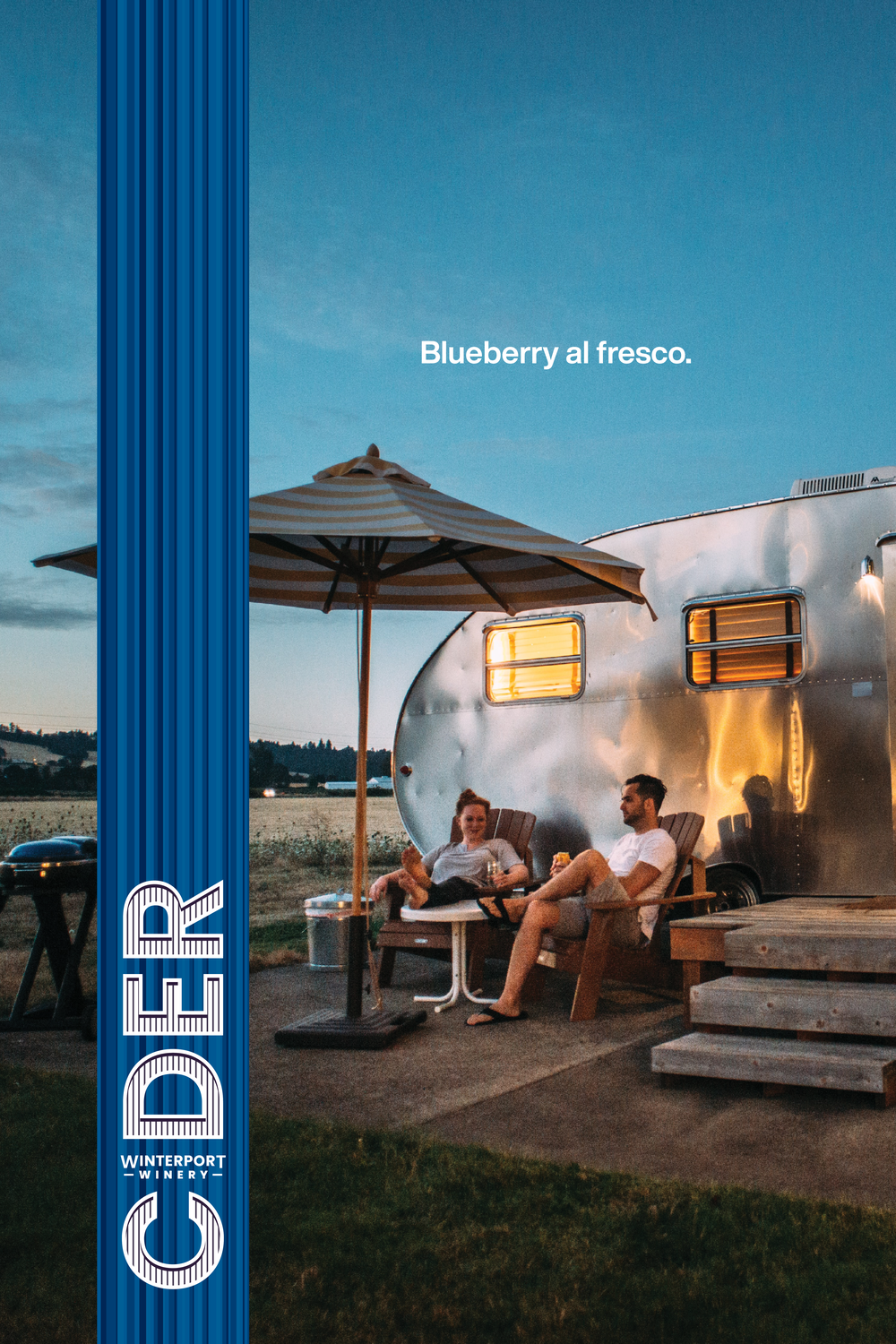 Two people relax outdoors by a vintage silver camper trailer, enjoying drinks under a striped umbrella during sunset. The scene captures a cozy, al fresco atmosphere, highlighting the enjoyment of nature and outdoor living.