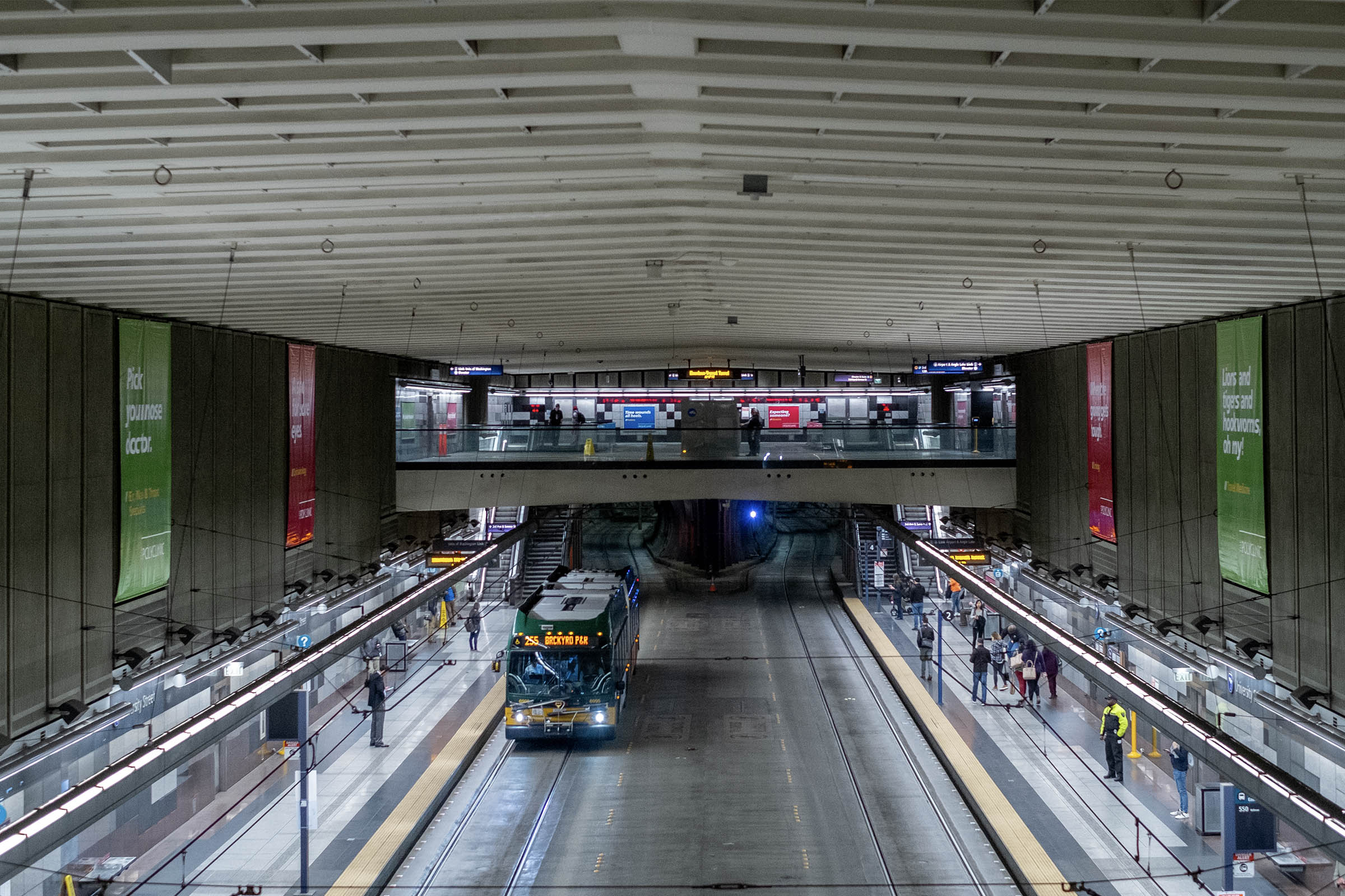 Urban transit station interior featuring a bus arriving at a platform, with passengers waiting and illuminated signage. The modern architectural design includes high ceilings and multiple levels.