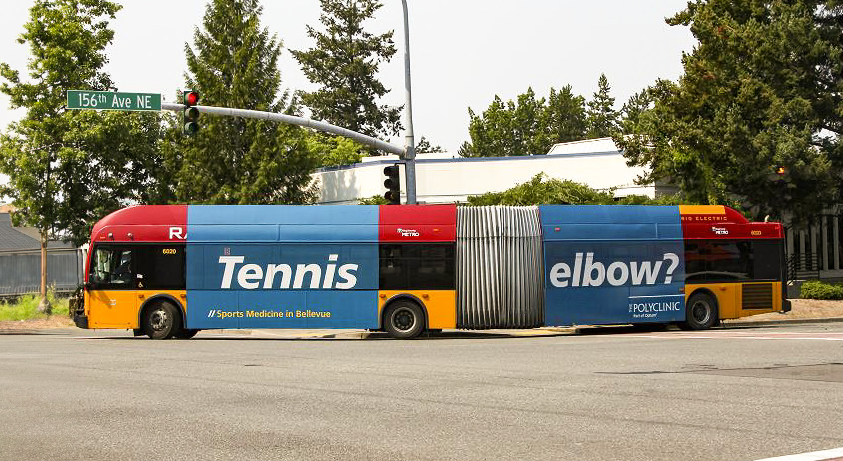 Blue public bus with a promotional wrap featuring the text "Tennis elbow" at a traffic light, surrounded by green trees.