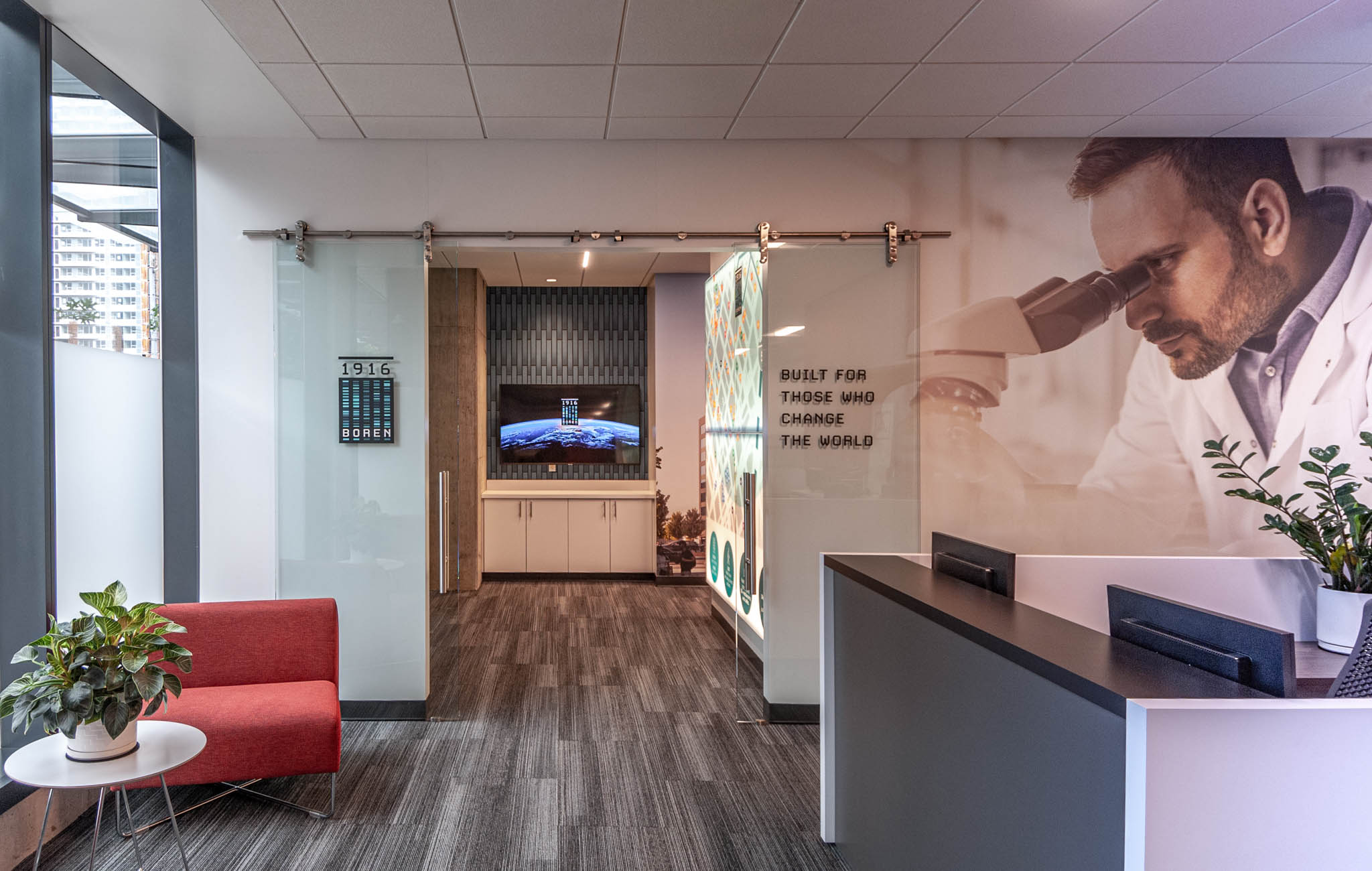 Modern office reception area featuring a sleek front desk, red seating, and a glass partition leading to a waiting area with a television display. The design incorporates contemporary decor and wood flooring, creating a welcoming atmosphere.