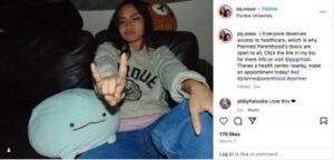 Young woman making a "rock on" hand gesture while sitting on a couch with plush toys, promoting healthcare access and Planned Parenthood services at Purdue University.