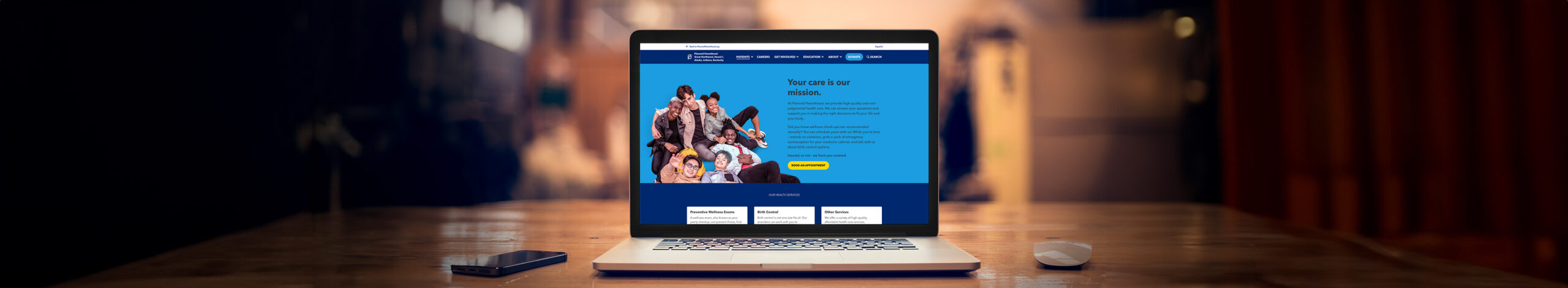 Laptop displaying a healthcare website with a blue background and diverse group of people, emphasizing the message "Your care is our mission." The setting features a wooden table and a smartphone beside the laptop.