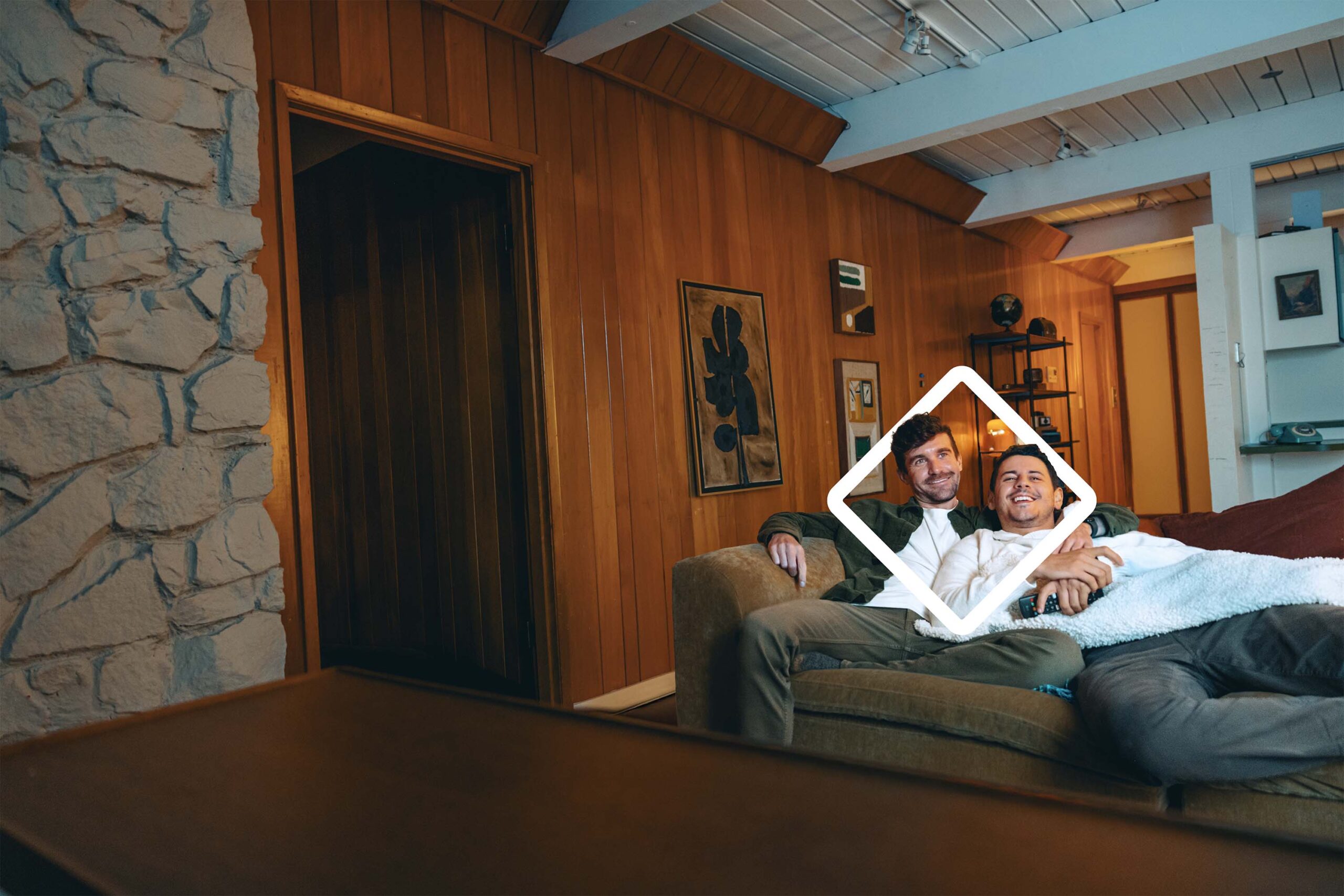 Two smiling men sitting on a couch in a cozy wooden-paneled living room, surrounded by modern art and decorative elements. The warm atmosphere is enhanced by natural lighting and stylish interior design.