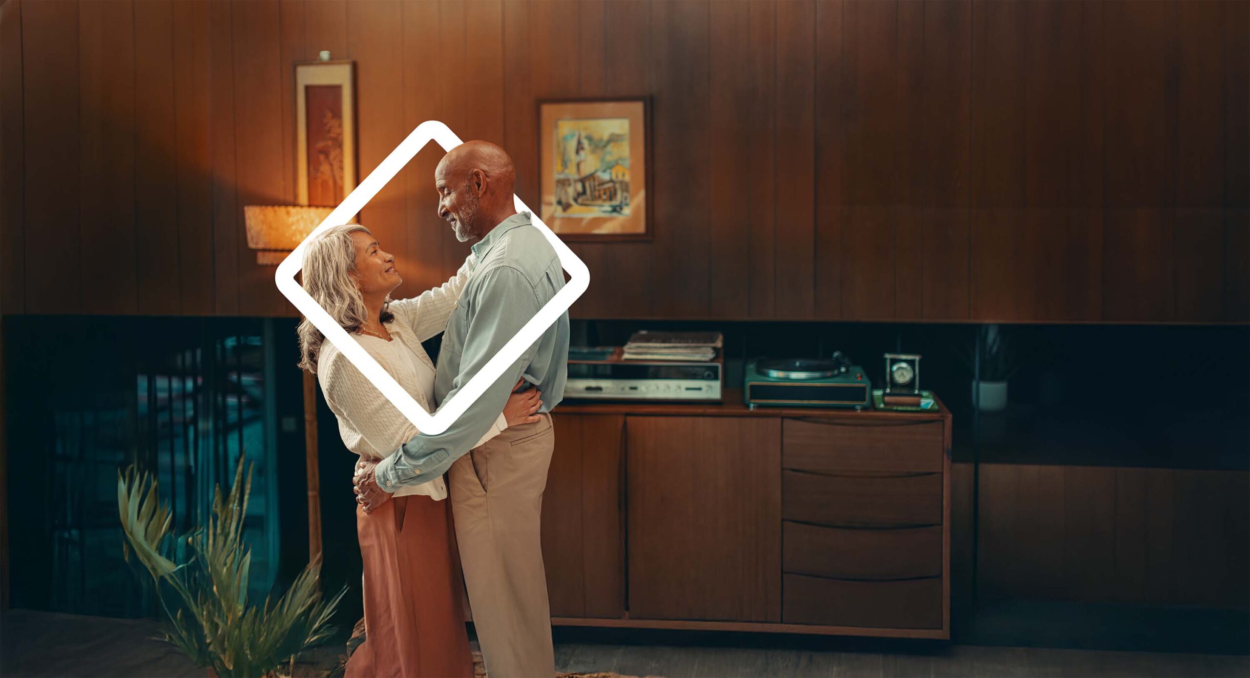 Elderly couple embracing in a cozy, vintage living room, showcasing warmth and affection. The room features retro decor, including a record player and artwork, highlighting a nostalgic atmosphere.