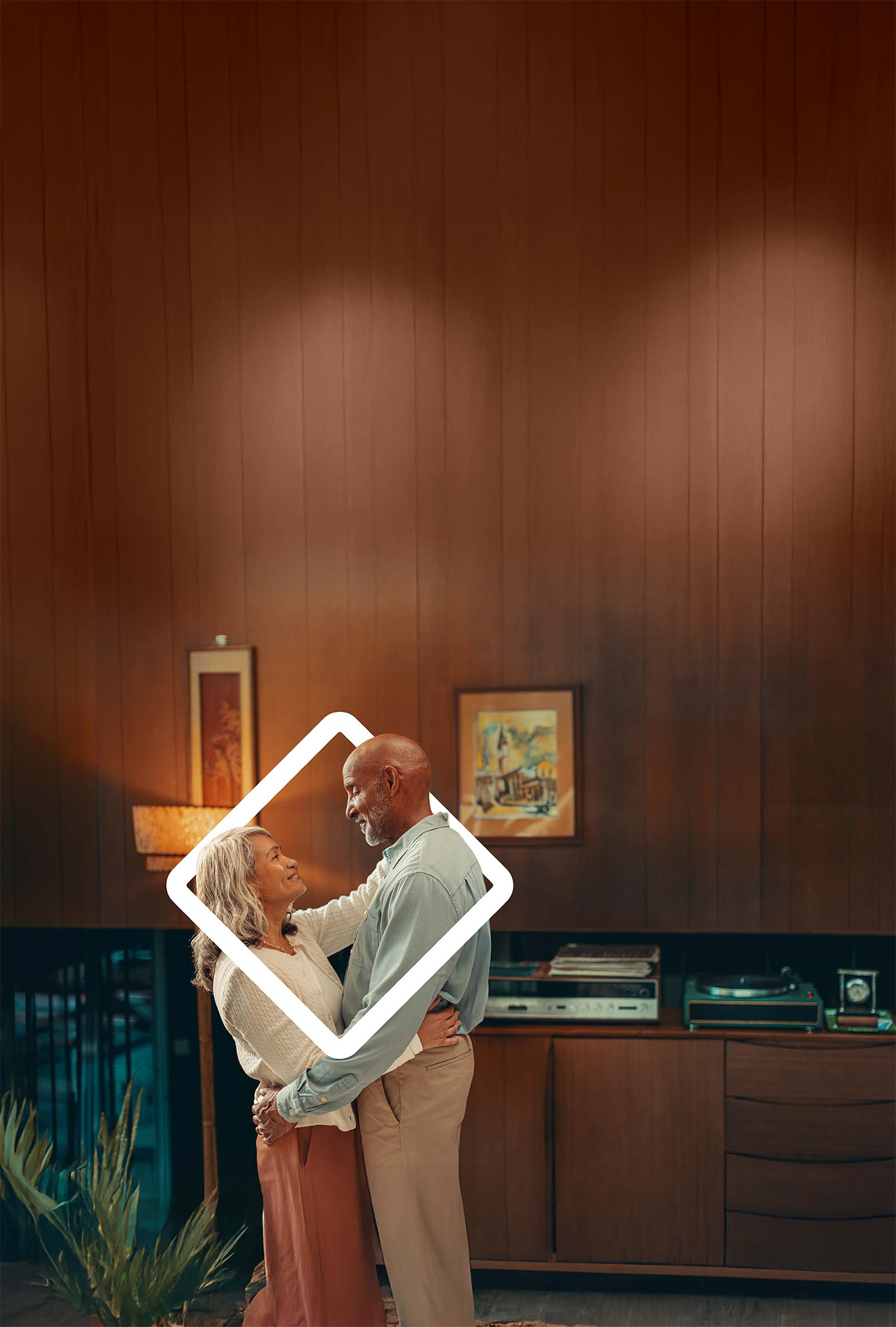 Couple embracing and smiling in a cozy, warmly lit living room with wooden walls and framed artwork, showcasing a moment of love and connection.
