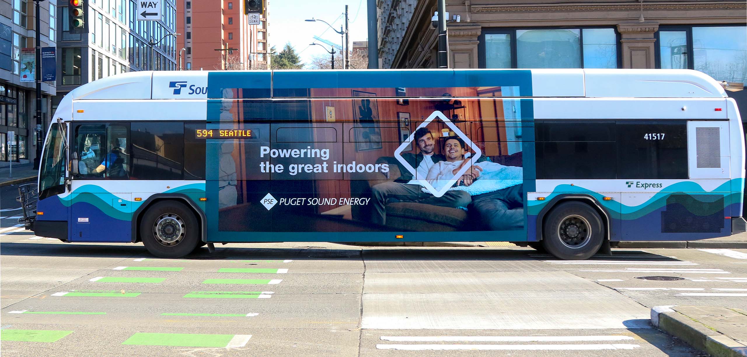 Bus advertisement for Puget Sound Energy in Seattle featuring two men relaxing indoors on a couch with the text "Powering the great indoors."