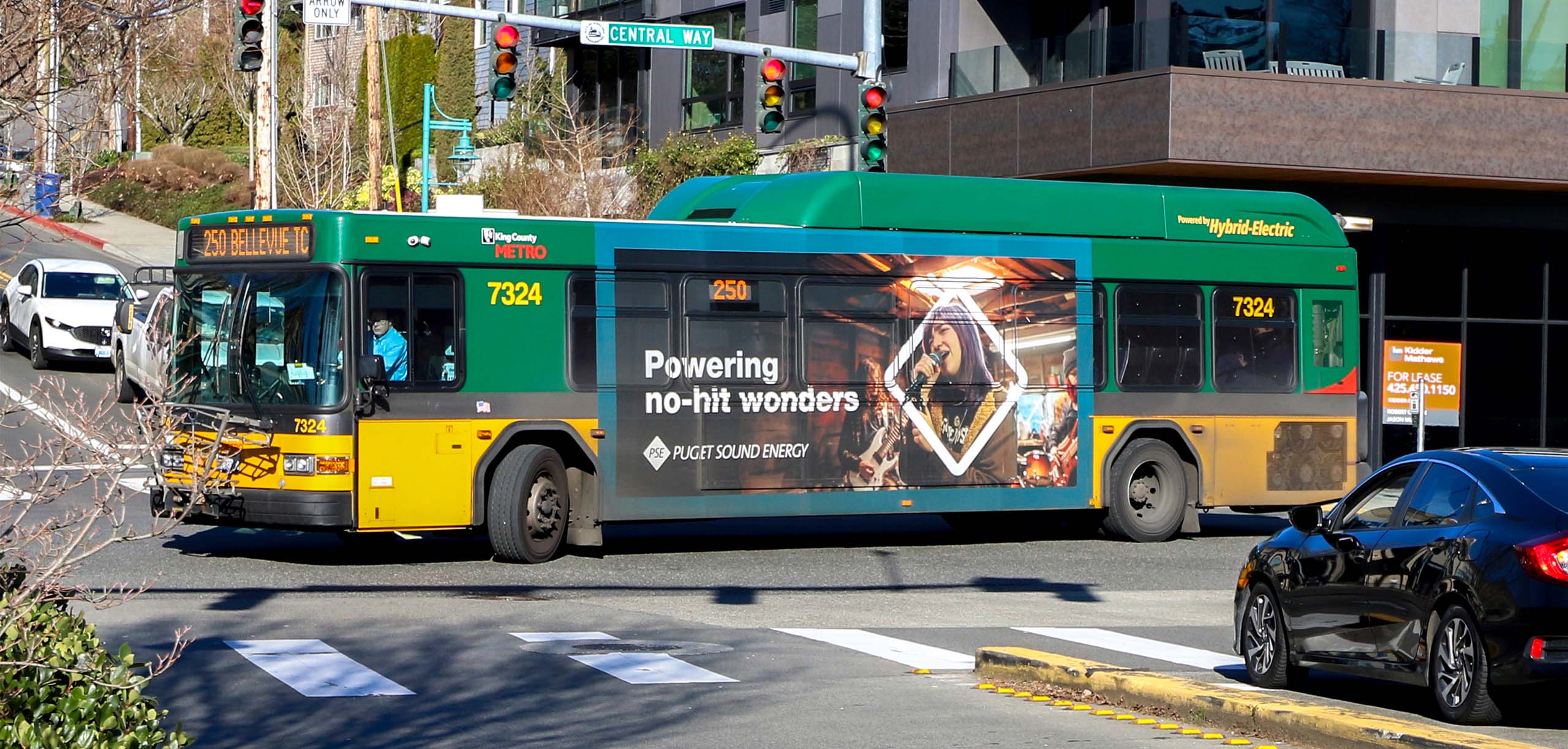Metro bus 7324 displaying an advertisement for Puget Sound Energy, featuring the slogan "Powering no-hit wonders" and promoting route 250, stopped at a traffic light on Central Way.
