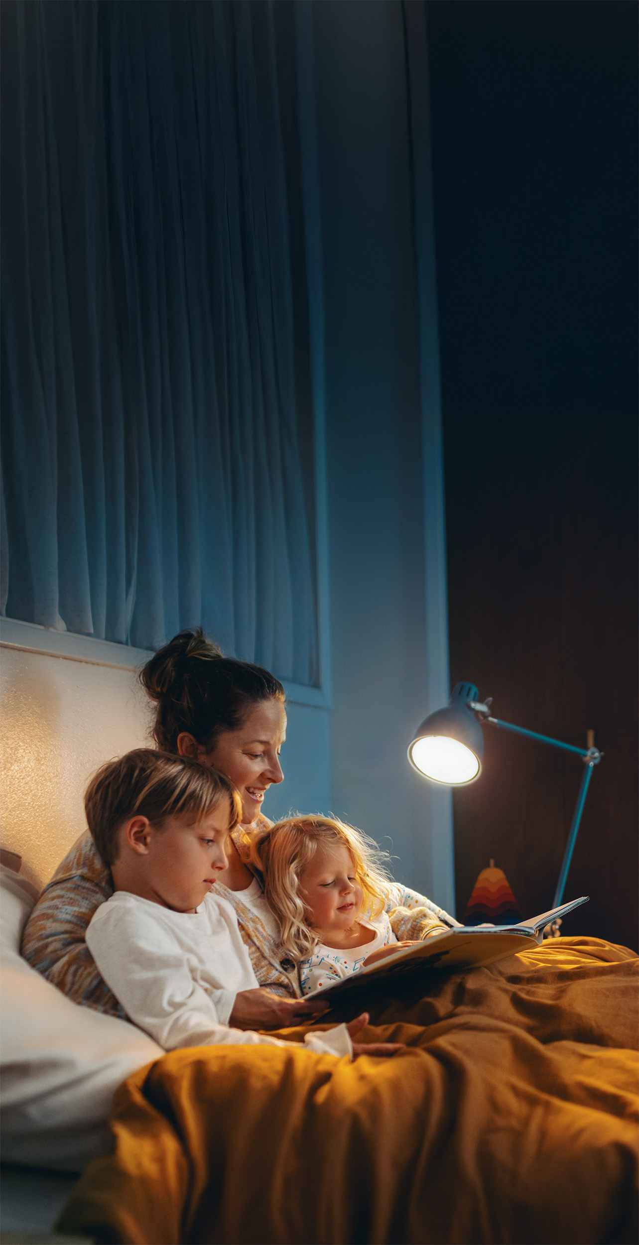 A mother sitting on a bed with two children, reading a story together in a cozy, softly lit bedroom. The atmosphere is warm and inviting, highlighting family bonding time.