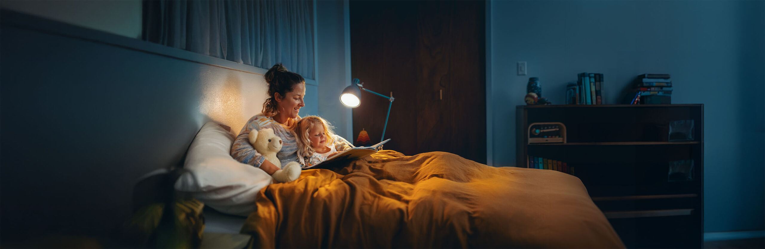 A mother reading a bedtime story to her young daughter in a cozy bedroom, illuminated by a bedside lamp, with a teddy bear resting beside them.