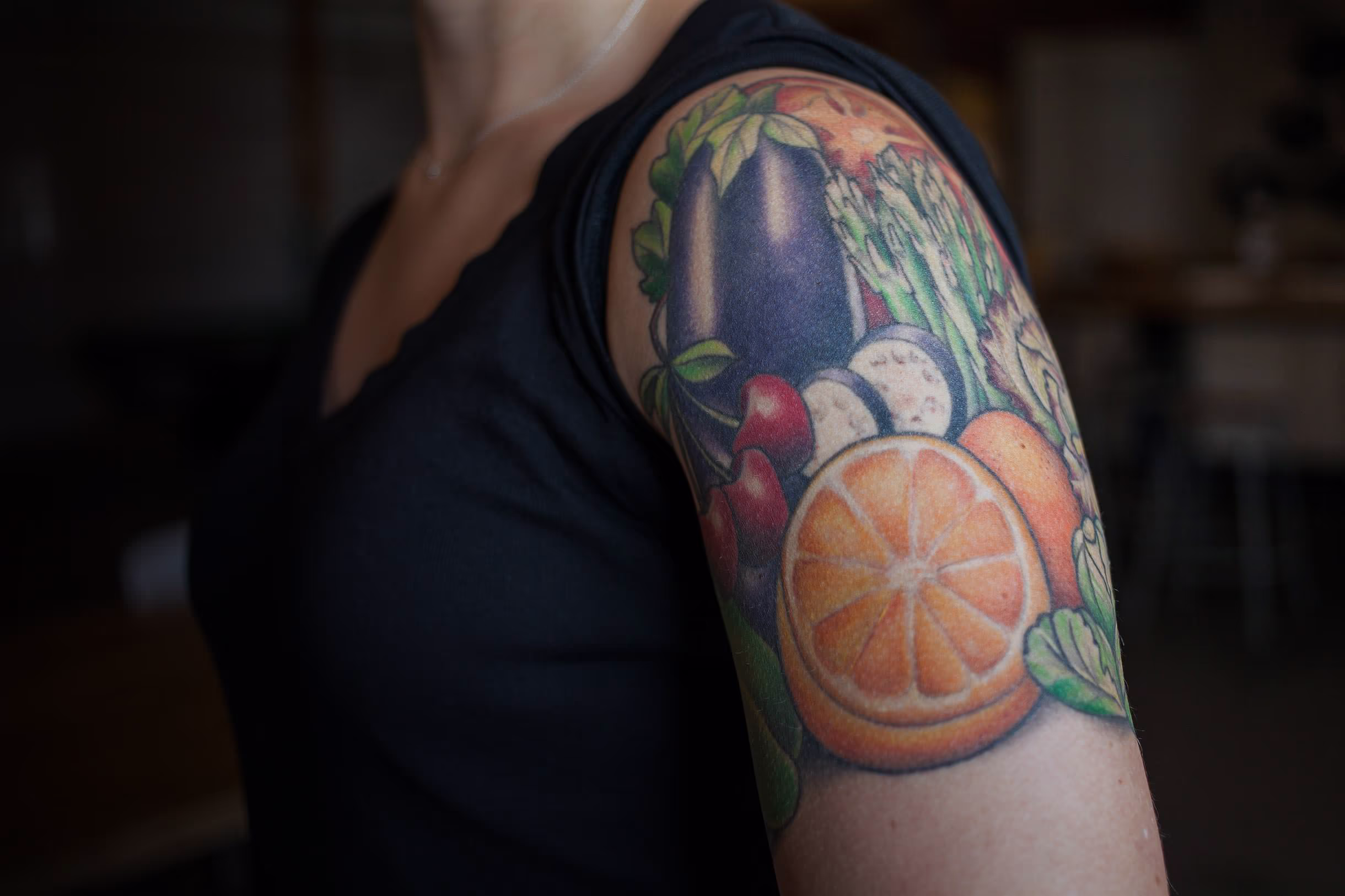 Colorful vegetable and fruit tattoo design on a woman's upper arm, featuring an orange, eggplant, asparagus, red peppers, and mushrooms, showcasing a vibrant and artistic representation of healthy food.