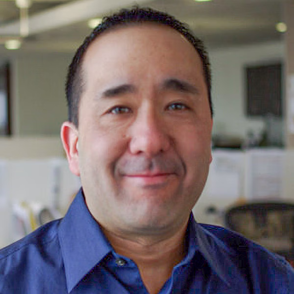 Professional headshot of a man with short black hair, wearing a blue collared shirt, smiling confidently in an office environment with blurred workstations in the background.
