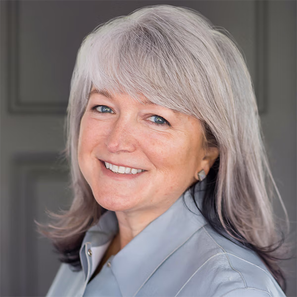 Smiling middle-aged woman with long, gray hair wearing a light blue blouse, posing in a professional setting.