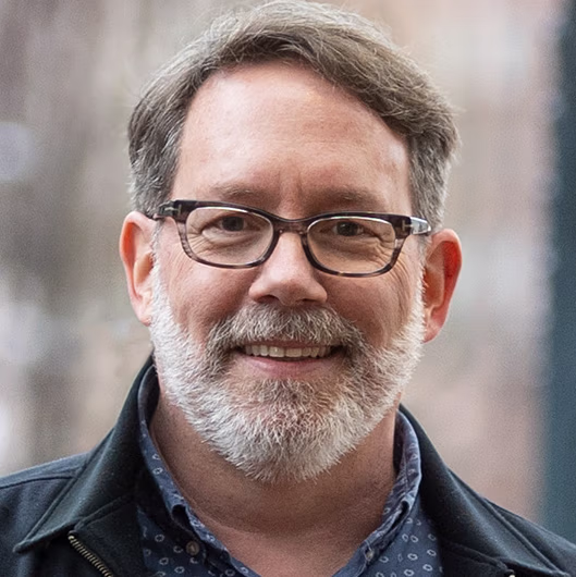 Smiling middle-aged man with glasses and a beard, wearing a dark jacket over a blue patterned shirt, standing against a blurred urban background.